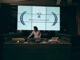 © Maximilian Röder || wwwmaxroedercom DJ behind a glass booth mixing music as a large screen displays '#beproud of yourself' with trophy and laurel symbols in a club setting