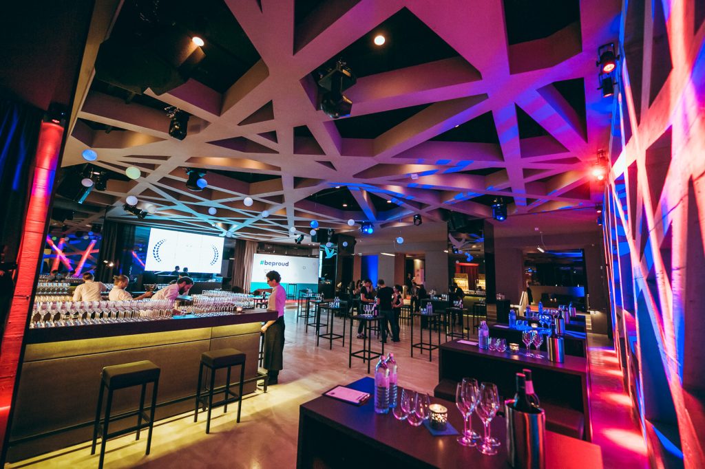 © Maximilian Röder || wwwmaxroedercom Bar area at a modern event space with a geometric ceiling, colorful pink/blue lighting, and rows of wine glasses on the counter