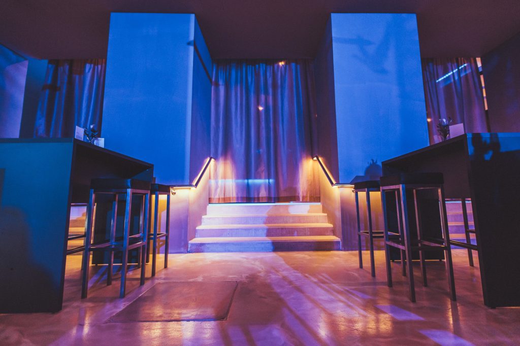 © Sergiu Andrés Dimly lit nightclub interior with blue lighting, center stairs, and high tables along the sides.