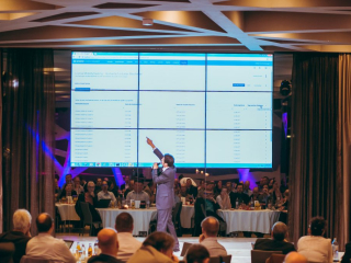 © Maximilian Röder || wwwmaxroedercom Male presenter in a gray suit speaks into a microphone on stage beside a large multi-panel screen displaying a data dashboard; audience sits at round tables below.