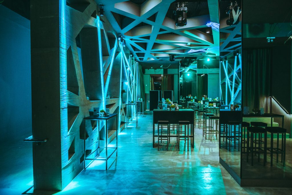 Fachvortrag Modern event venue with teal lighting, geometric white ceiling, and tall tables set for guests in a long, open hall.