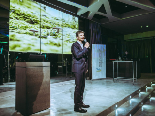 Fachvortrag Man in a suit speaks into a microphone on a stage with a large video wall behind him and a banner to the side.