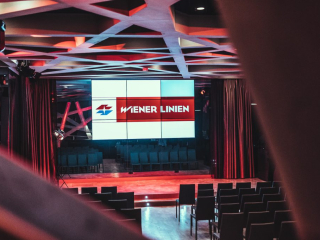 Palais_Wertheim_Fachvortrag_Wiener-Linien-16 Empty conference stage with a six-screen LED display showing the Wiener Linien logo in red and blue, lit by red stage lights.