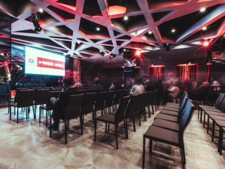 Palais_Wertheim_Fachvortrag_Wiener-Linien-26 Large conference room with rows of chairs facing a stage and a bright LED screen, red and blue stage lighting creating a modern, energetic atmosphere