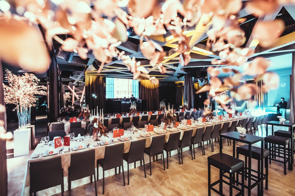 Palais_Wertheim_Galadinner_Speedinvest-2 Banquet hall set for a formal dinner with long rows of tables, red place cards, candle centerpieces, and a stage with performers in the background under dramatic ceiling lights.