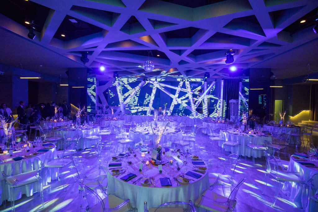 Palais Wertheim Elegant banquet hall set for a formal dinner with round tables, clear chairs, white linens, and blue-purple lighting; a geometric ceiling and stage backdrop dominate the space.