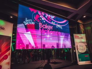 Palais_Wertheim_Jahresfeier_BakerMcKenzie_-73 Stage setup for Baker McKenzie’s 15th anniversary with a large, colorful LED wall displaying 'Baker McKenzie' and 'Happy Birthday' graphics, plus event banners nearby.