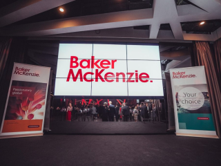 Palais_Wertheim_Jahresfeier_BakerMcKenzie_-97 Large screen displays the Baker McKenzie logo in red, flanked by event banners; attendees mingle in a conference venue behind.