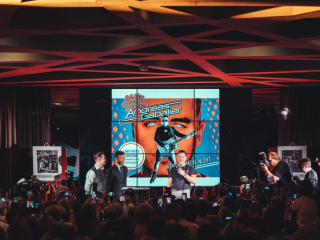 Palais_Wertheim_Konzert_Andreas-Gabalier-32 Man on stage singing into a mic in front of a giant poster of a guitarist, as an audience films with their phones.