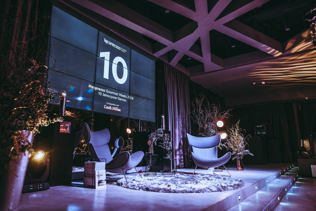 Special Event by Nespresso Stage setup with a large screen showing '10' for Nespresso Gourmet Weeks 2018, two curved blue chairs, and winter decor with faux snow and branches.