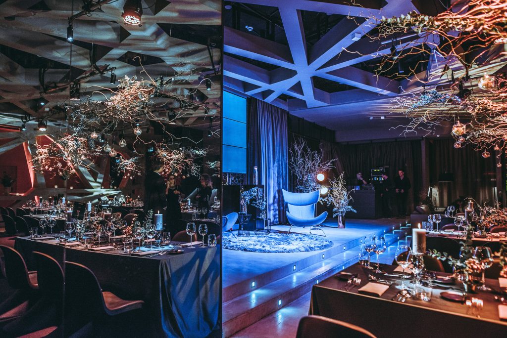 Special Event by Nespresso Banquet hall setup with tables, glasses, and candles; a lit blue-stage seating area with a curved blue chair and decorative bare branches to the right.