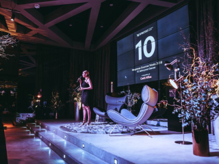 Special Event by Nespresso Female presenter in a black dress speaks into a microphone on stage beside a large screen showing '10' at a formal event.