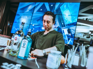 Palais Wertheim | Coca Cola Bartender in a green shirt behind a bar, arranging bottles with a blue-lit multi-panel screen in the background.