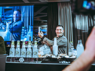 Palais Wertheim | Coca Cola Man at a bar taking a selfie with his phone, bottles and bowls of chips on the counter, a large screen behind him.