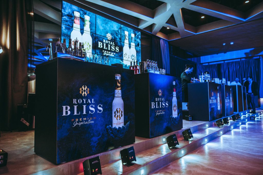 Palais Wertheim | Coca Cola Row of black bar stations with Royal Bliss branding and bottles, blue lighting, large screen showing Royal Bliss ads in the background.