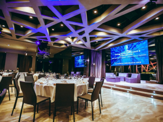 © Maximilian Röder || wwwmaxroedercom Elegant conference hall set for a formal luncheon with round tables, white linens, and a stage with a blue presentation screen.