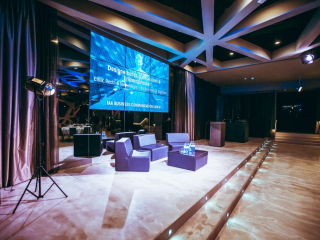 © Maximilian Röder || wwwmaxroedercom A modern event stage with a large blue digital screen, purple seating area, and a row of lit steps leading to a dark back area; water bottles on a small table in front of the chairs.