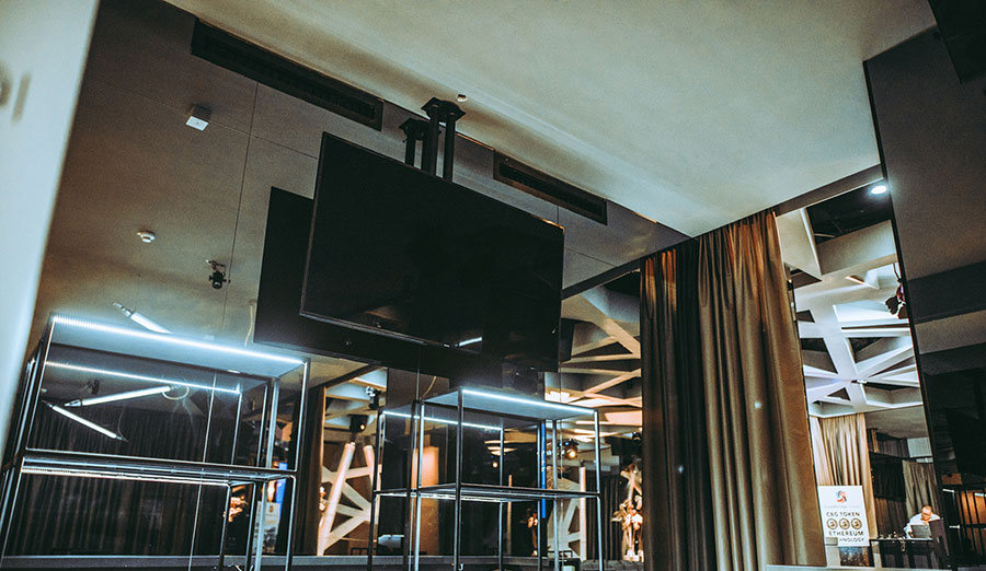 Large flat-screen TV mounted above glass-and-metal shelving in a modern lobby with blue neon lighting.