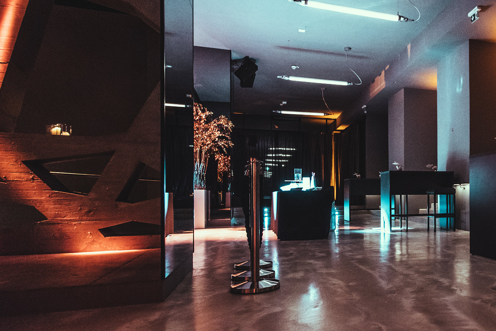 Dim, modern lounge or club interior with blue and orange lighting, a polished concrete floor, and high tables in the background.