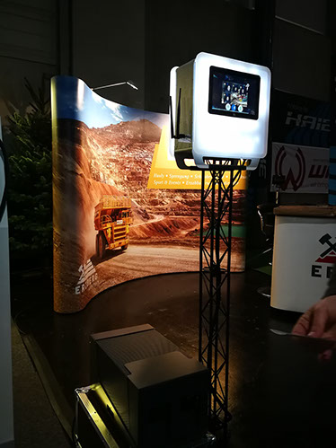 palais-wertheim-fotobox-1 - Palais Wertheim Eventlocation Wien Touchscreen display mounted on a tall stand at an expo booth, with a desert quarry backdrop and a yellow truck image behind it.