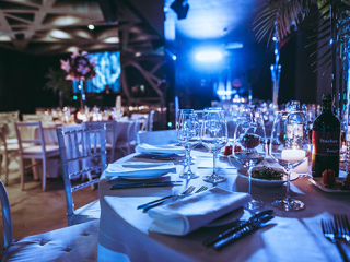 Banquet table set for a formal event with white linens, glassware, candles, and blue stage lighting.