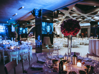 Formal banquet hall with round tables, white chairs, and tall pink flower centerpieces under blue lighting in a modern venue.
