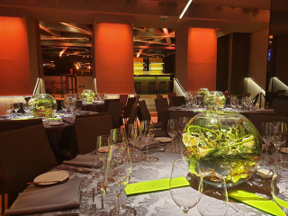 Elegant banquet hall with round tables set for a formal event, water glasses and napkins arranged around a large glass centerpiece containing greenery and flowers in the foreground.