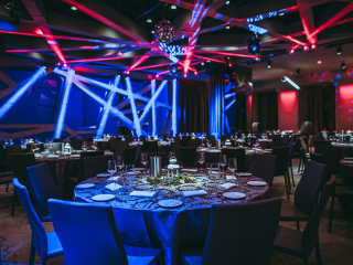Banquet hall with round tables set for dinner; blue and red stage lighting creates a dramatic atmosphere.