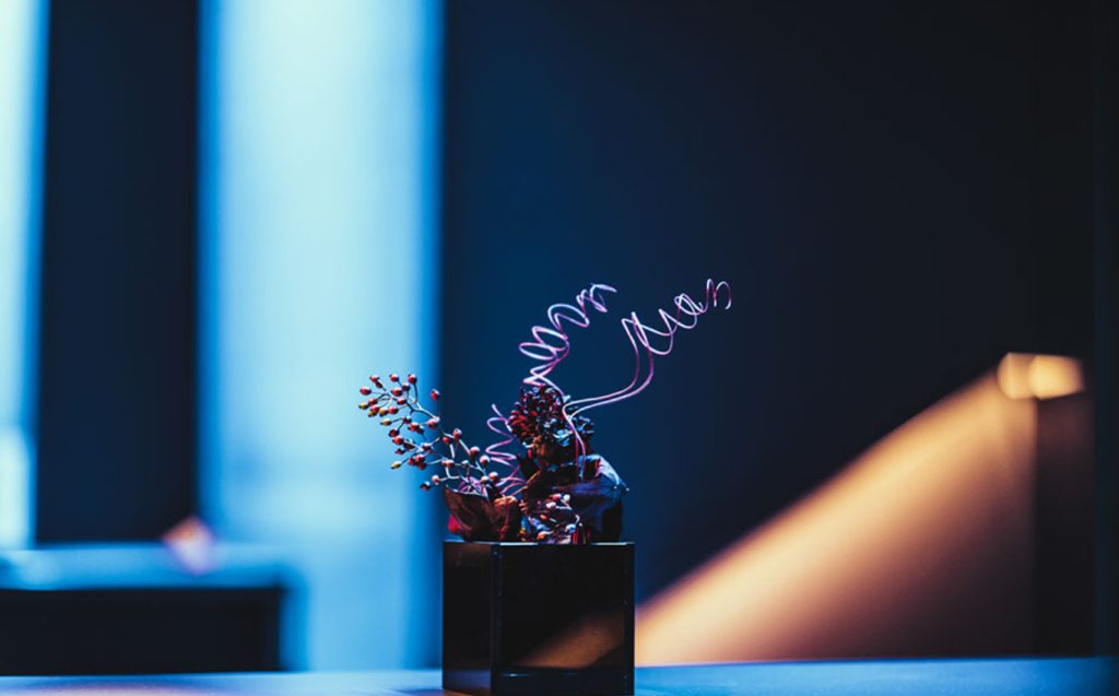 Small black square vase with red berry branches and pink curly stems on a blue-lit table surface to create a moody centerpiece.