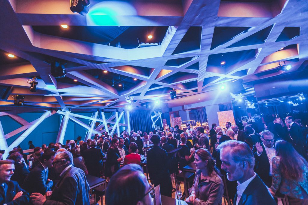 © Sergiu Andrés Professionals mingle at a blue-lit corporate reception in a futuristic venue with white crisscross ceiling beams.