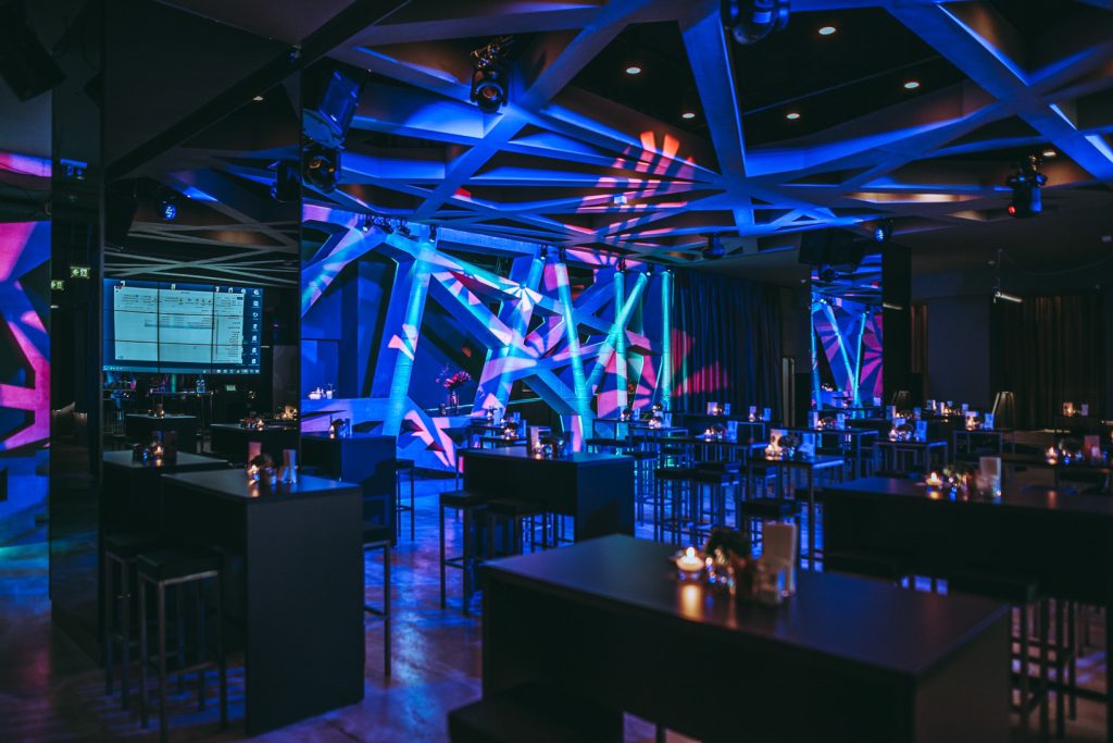 Fachvortrag Stylish event venue with blue and purple lighting, angular lattice beams, and candlelit high tables scattered throughout.