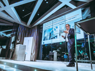 Fachvortrag Male presenter in a suit giving a talk on a stage with a large screen showing slides about a knowledge center; audience visible behind glass panels.