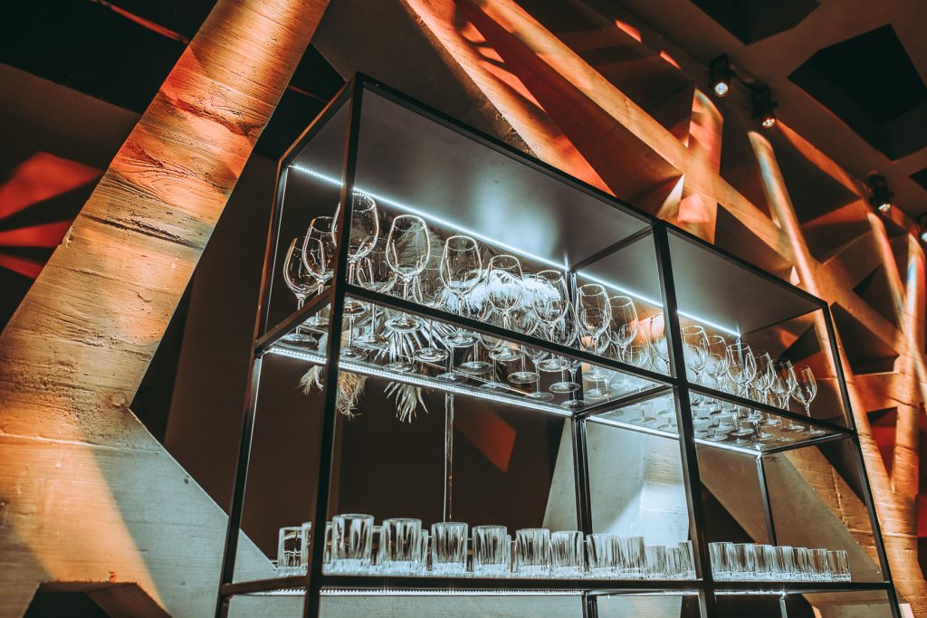 Palais Wertheim | Galadinner Glassware display on metal shelves illuminated by blue LED lighting in a modern bar setting with warm orange ambient light behind geometry of the walls.