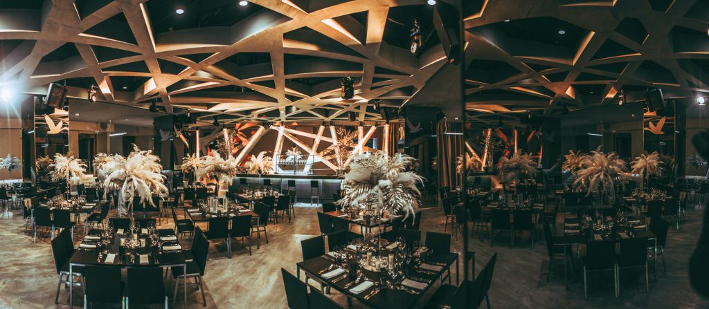 Palais Wertheim | Galadinner Elegant banquet hall with geometric wood ceiling, round tables, and palm centerpieces, stage lights in the background.