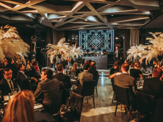 Palais Wertheim | Galadinner Guests in suits at a formal banquet, facing a stage with a speaker and geometric backdrop screen.