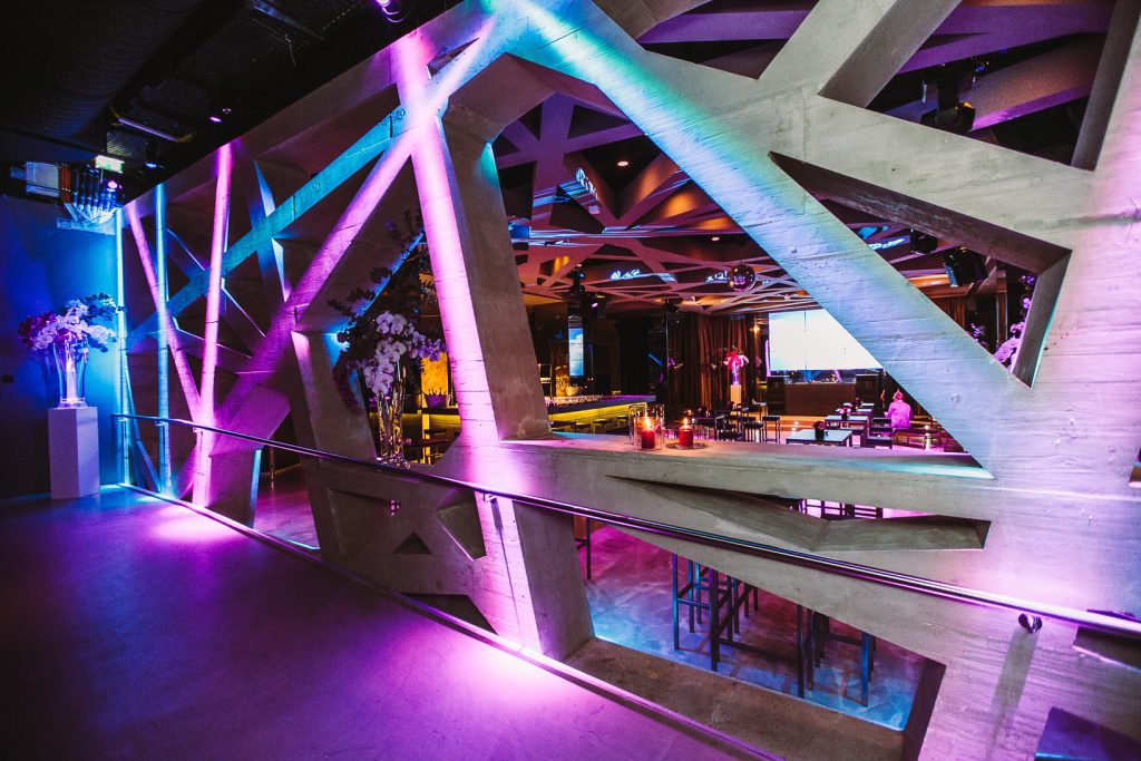 Palais Wertheim | Geburtstagsfest Industrial club interior with large diagonal concrete beams lit in blue and pink, revealing a seating area with candles and flowers through the framework.