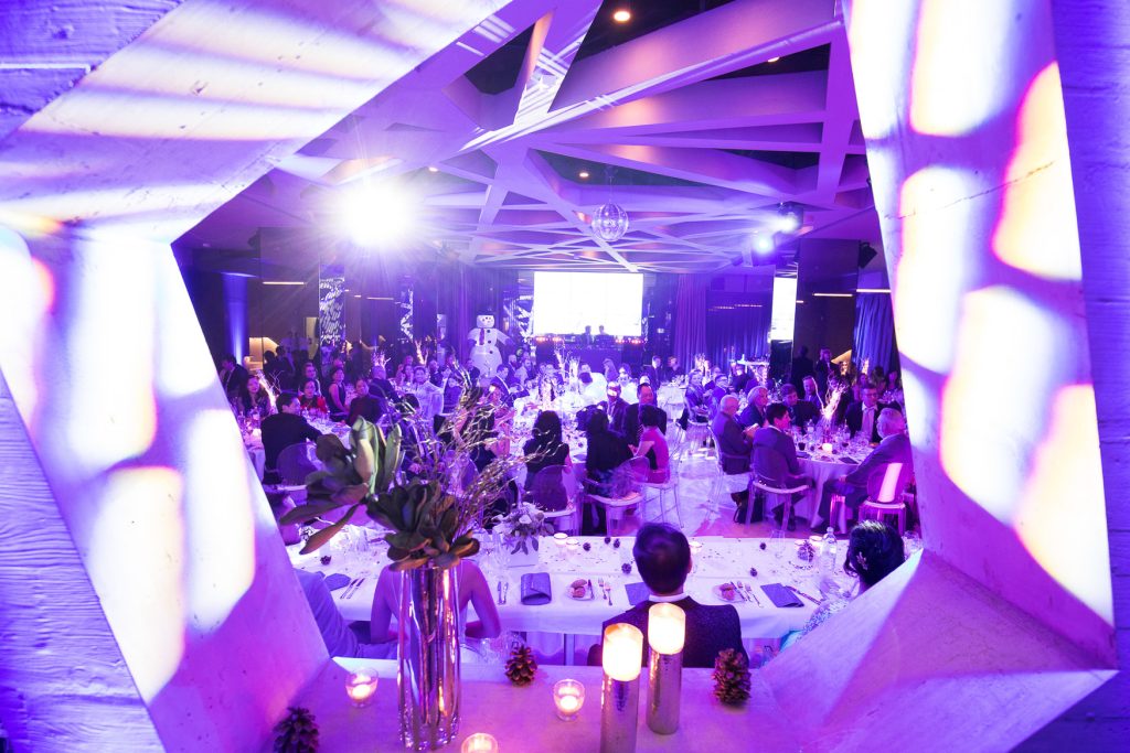 Palais Wertheim Banquet hall scene seen through geometric frame, guests seated at long tables with candles and floral centerpieces in purple lighting.