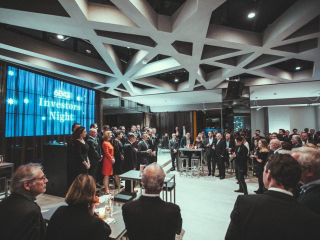 © Sergiu Andrés Crowd of professionals in suits at a corporate Investors Night, with a blue screen on the left reading 'Investors Night'.