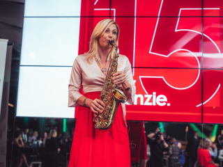 Palais_Wertheim_Jahresfeier_BakerMcKenzie_-64 Female saxophonist performing on a stage with a bright red digital backdrop behind her.