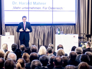 Palais Wertheim Speaker in a dark suit and purple tie on a stage, gesturing, with a large screen behind displaying text in German and a seated audience in front.