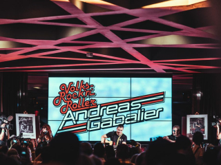 Palais_Wertheim_Konzert_Andreas-Gabalier-34 Live band performs on a stage with a large LED screen behind showing rock ’n’ roll graphics and the performer’s name; camera crews and audience in foreground.