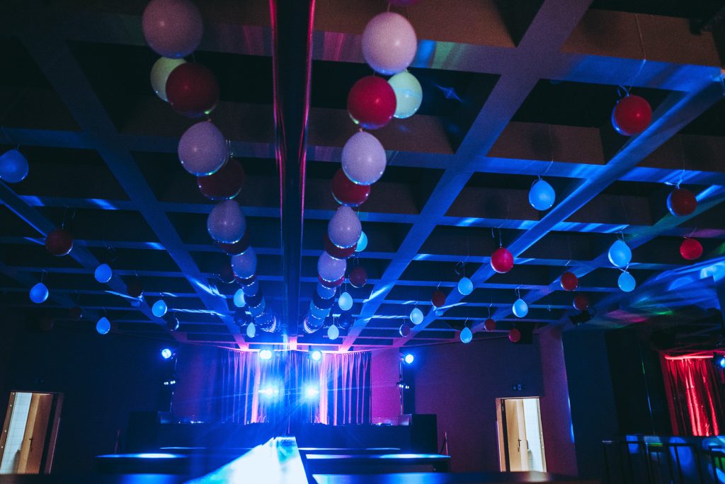 Palais_Wertheim_Schulball_Goethegymnasium-1 Balloon strands hanging from a grid ceiling over a dimly lit party space with blue/purple stage lights and a DJ booth in the background.