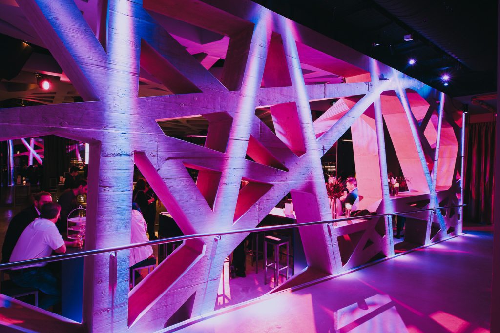 Palais Wertheim Geometric wooden lattice partition bathed in pink and purple lighting, with people seated at a bar in the background