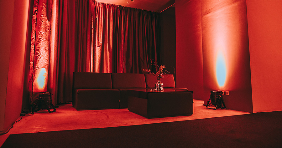 Red-lit lounge area with a black sectional sofa, matching ottoman, and a glass vase on the coffee table.