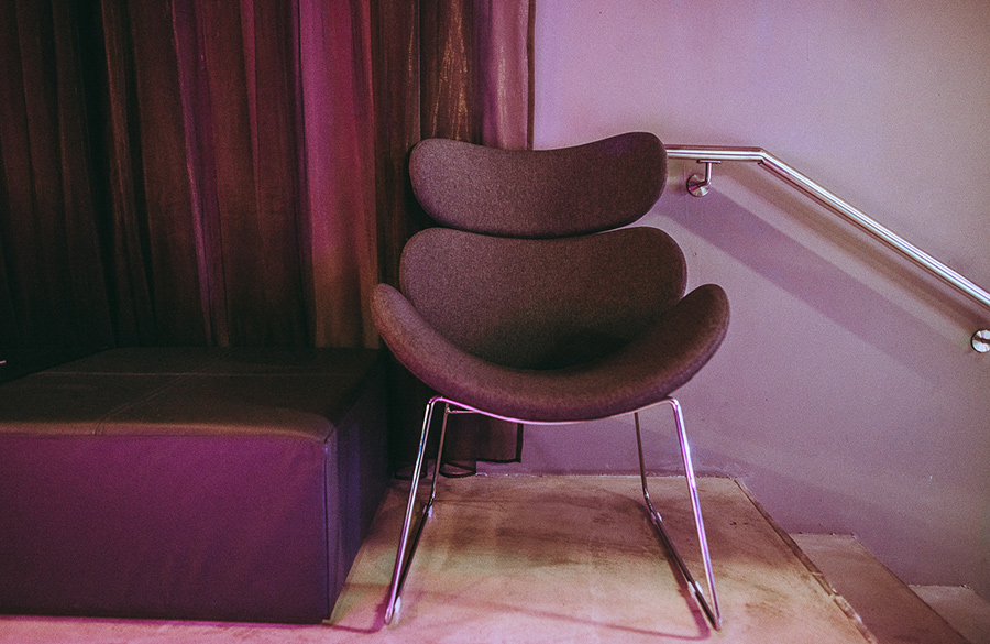Modern curved lounge chair with chrome legs in a purple-lit corner beside a purple bench.