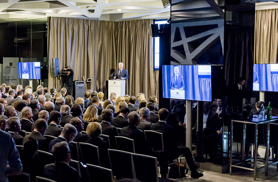 Speaker at a podium addressing a large seated audience in a conference room with video screens displaying the speaker nearby