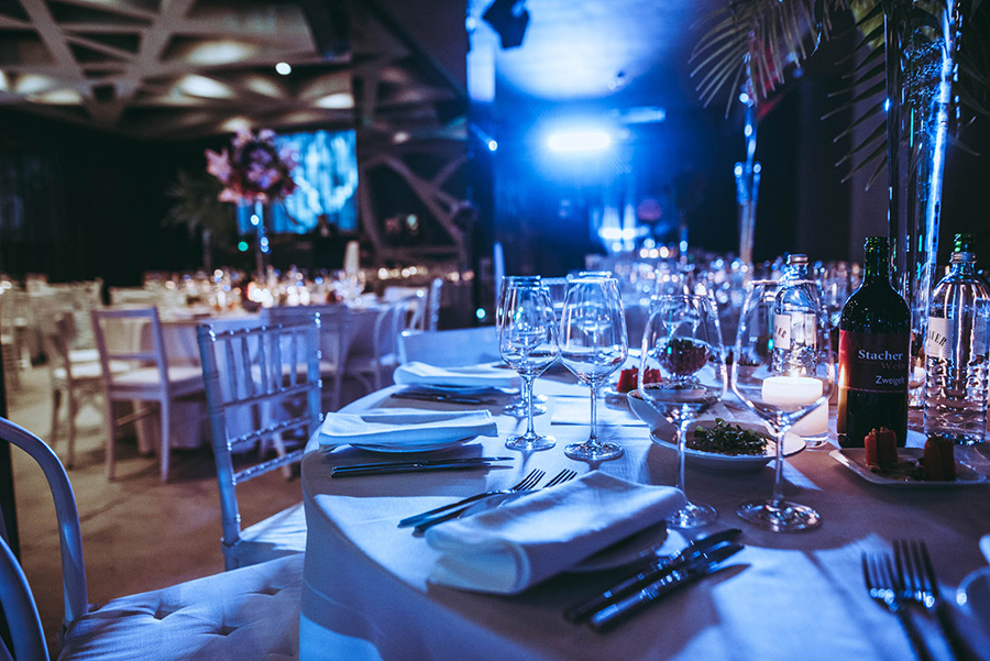 Elegant banquet table set for a formal dinner with white tablecloths, wine glasses, and bottles under blue lighting on stage