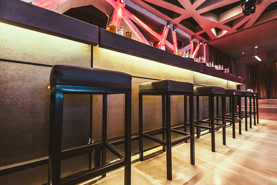 Long bar with black stools lined up along a glowing countertop in a modern lounge or club setting, bottles visible on the back rail.