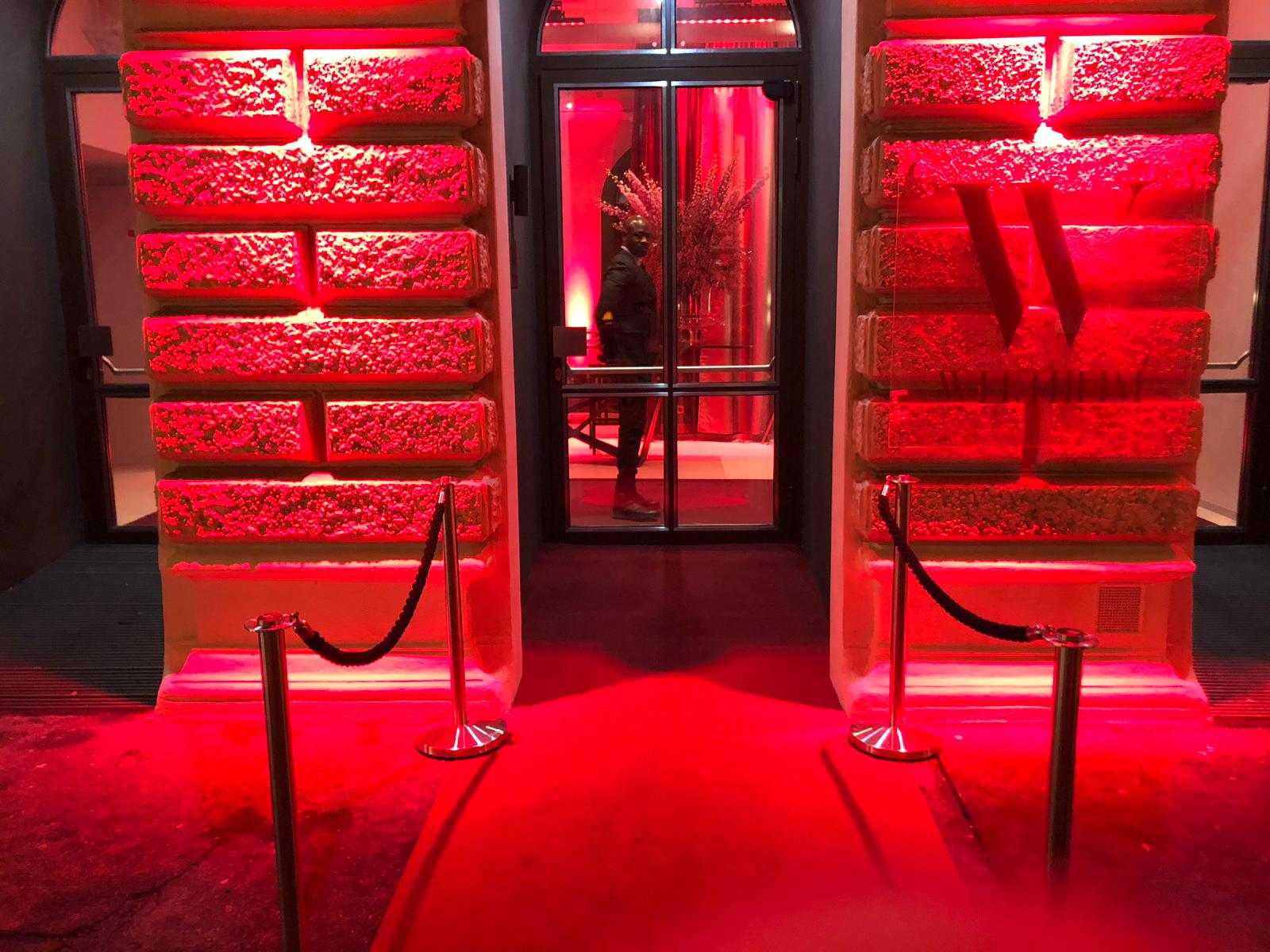 palais-wertheim_lichtanlage_foyer-fassade - Palais Wertheim Eventlocation Wien Red-lit entrance framed by textured stone columns, glass doors in the middle, with rope barriers on either side and a person inside near the door.",