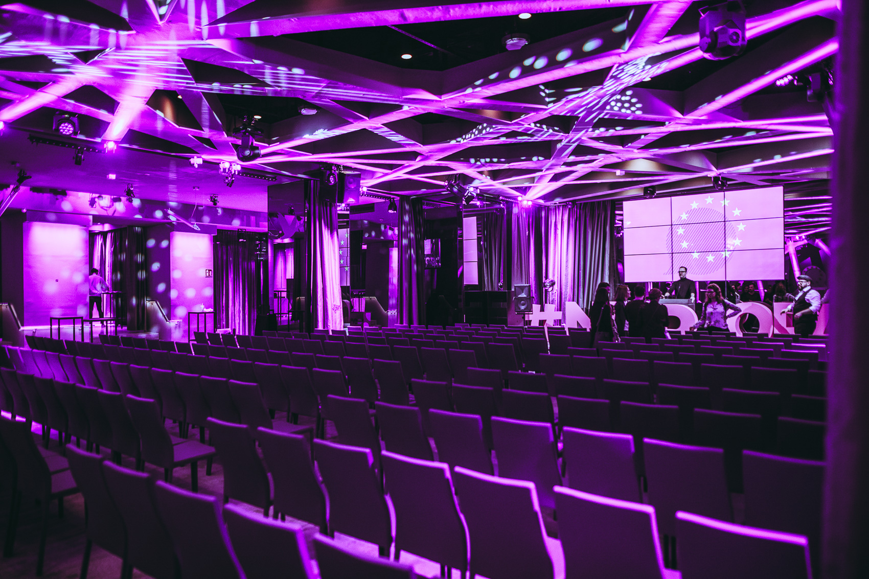 palais-wertheim_lichtanlage_wertheim-saal - Palais Wertheim Eventlocation Wien Conference hall with purple lighting, empty rows of chairs, and a stage with a large screen displaying a circular EU-like emblem.